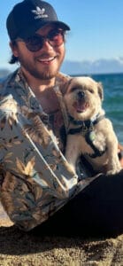 A person wearing sunglasses, a black cap, and a floral shirt sits on a beach holding a small dog with a harness. The sea and sky are visible in the background.
