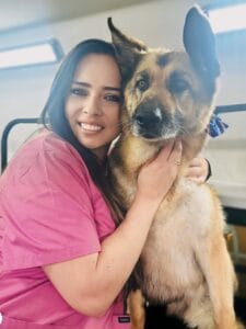 A woman in a pink scrub top, likely a dog groomer, hugs a German Shepherd as they look at the camera inside a well-lit room.