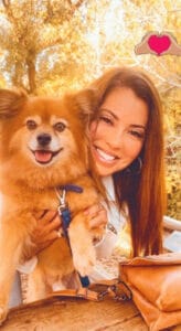 A woman smiles while hugging a fluffy brown dog outdoors, with trees in the background and a tan purse on the table in front.