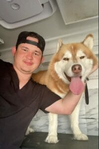 A man wearing a black cap and black shirt, clearly a pet lover, poses for a photo with a tan and white dog with its tongue out, standing beside him.