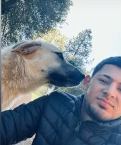 A pet lover in a black jacket takes a selfie outdoors while a large light-colored dog sniffs his ear. Trees are visible in the background.